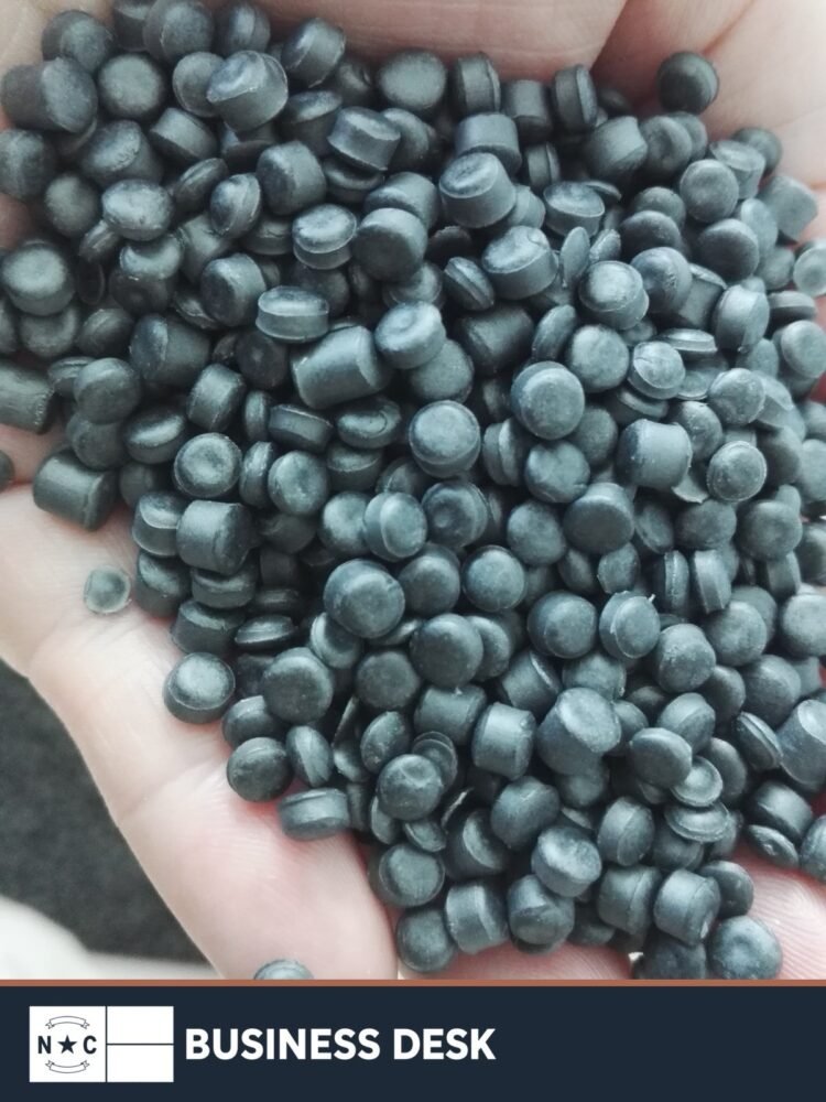 Plastic injection-molding pellets in a person's hand, shown as manufacturing context, above the NC Business Desk footer.