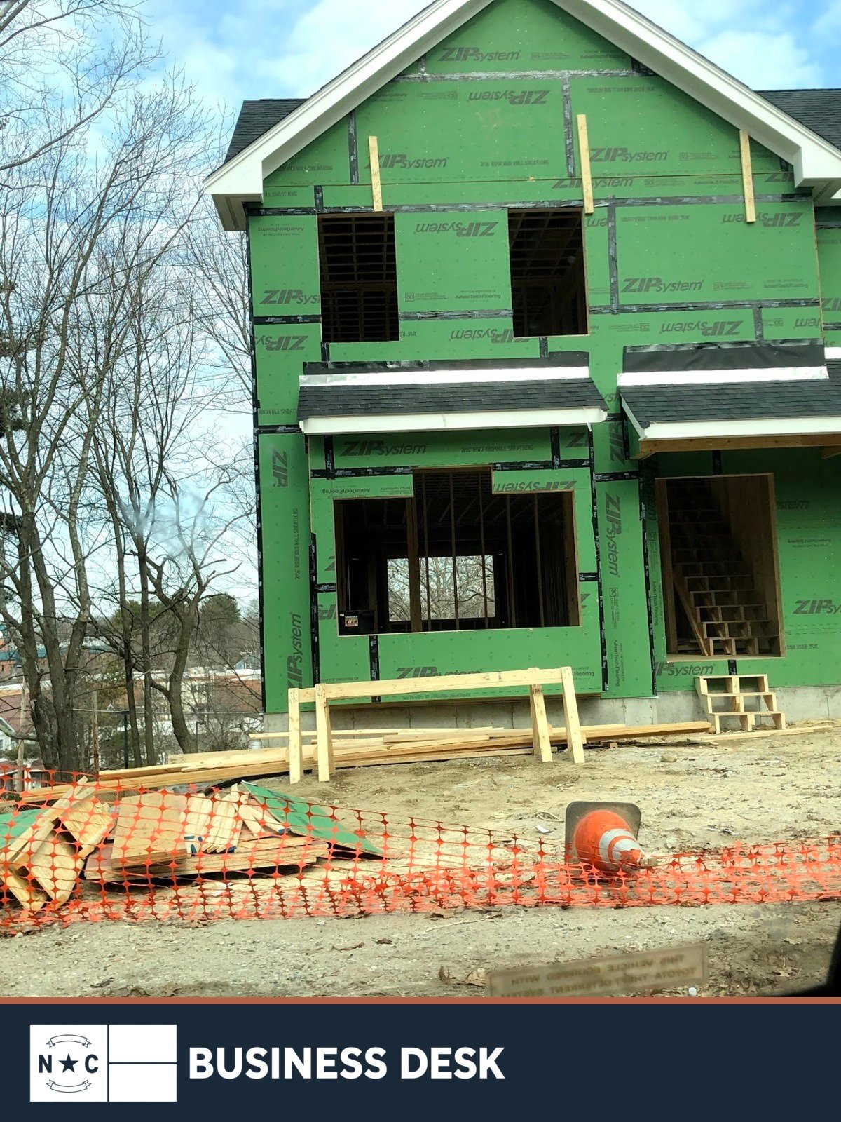 A single-family house under construction with green sheathing, lumber and safety fencing above the NC Business Desk footer.