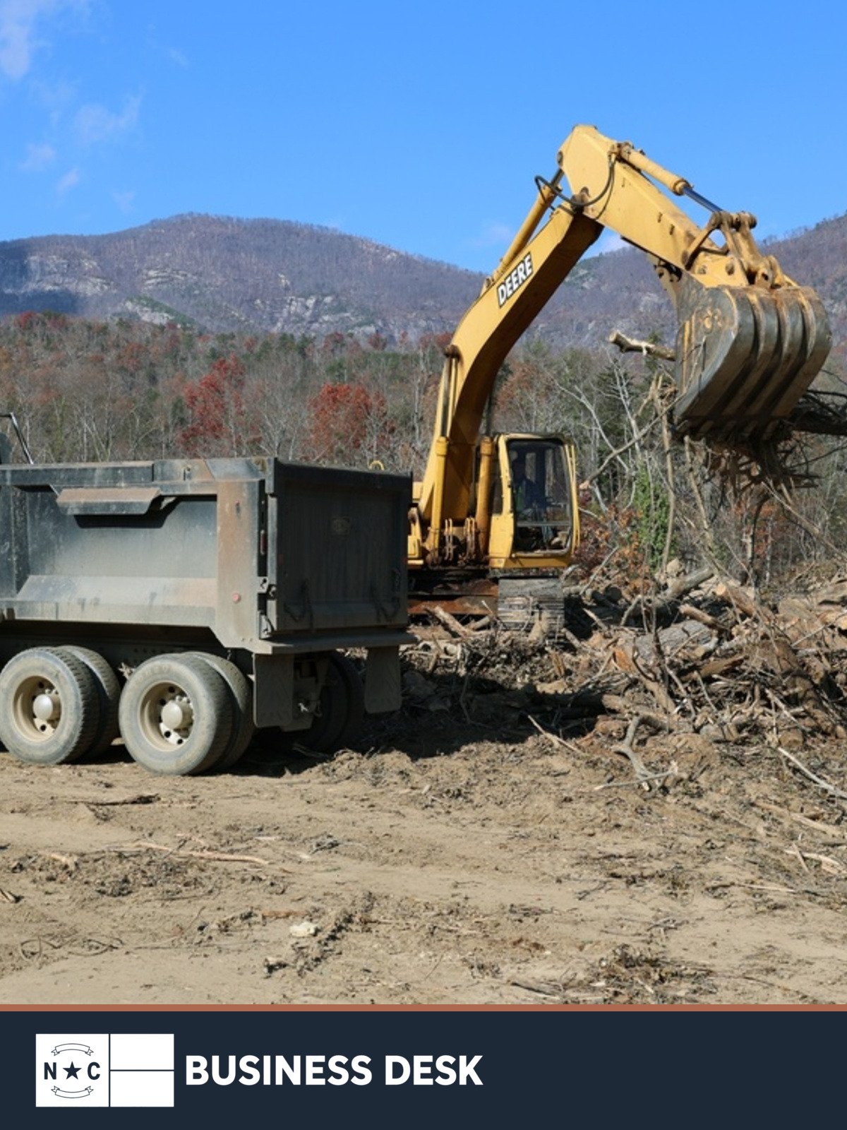 A tracked excavator loads Hurricane Helene vegetation debris into a dump truck at Morse Park in Lake Lure, North Carolina, with mountains in the background.