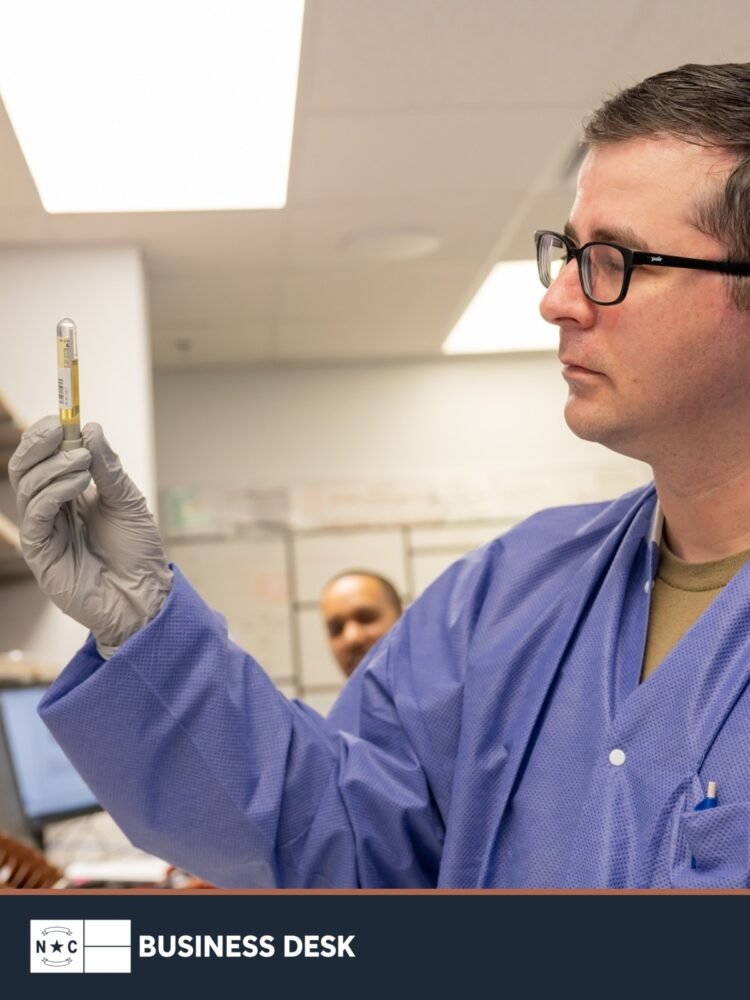 A medical laboratory technician examines a specimen in a clinical laboratory, shown with the NC Business Desk footer.