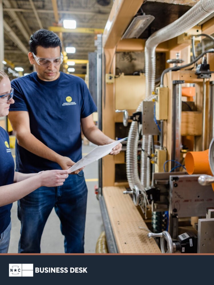 Two Pella factory employees review production documents beside manufacturing equipment above the NC Business Desk footer.